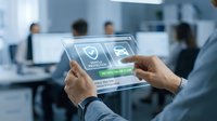 Businessman using transparent digital tablet with security shield icons in modern office environment for cybersecurity and data protection concepts.