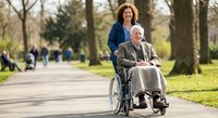 Caring middle-aged Caucasian woman pushing elderly Caucasian man in wheelchair through sunny park pathway. Healthcare assistance and mobility support concept.