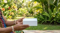 Woman with dark skin tone holding blank white business card in garden setting with lush green foliage and natural sunlight, suitable for branding, marketing, and corporate identity projects.