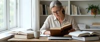Mature Caucasian woman with glasses reading book at home office desk surrounded by books and papers, focused on learning and research activities.