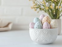 Pastel speckled Easter eggs in white ceramic bowl with geometric pattern, spring flowers in background on light surface. Minimal holiday composition.