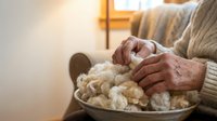 Elderly hands knitting with cream wool yarn in cozy home setting. Senior woman crafting handmade sweater. Retirement hobby and traditional needlework skills.