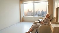 Middle-aged Caucasian woman in beige suit relaxing in modern office chair by window with city skyline view during golden hour for business lifestyle concepts.