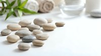 Smooth pebbles arranged on white surface with spa elements, towels and greenery in soft focus background for wellness and relaxation concepts.