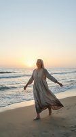 Joyful middle-aged Caucasian woman in flowing dress dancing on beach at sunset with arms outstretched, ocean waves and golden sky creating serene atmosphere.