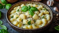 Creamy gnocchi pasta in ceramic bowl garnished with fresh basil leaves, black pepper, and parmesan cheese on rustic wooden background with garlic cloves.