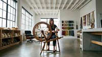 Young Asian woman artisan spinning wool on traditional wooden spinning wheel in bright modern textile studio workspace with colorful yarn samples displayed on walls.