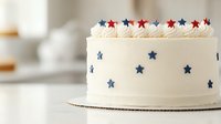 Patriotic white layer cake decorated with red and blue fondant stars and whipped cream piping along top edge, photographed on light background for holiday celebration.