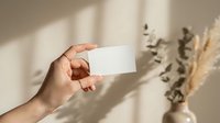 Hand holding blank white business card mockup with natural shadow overlay and dried flowers in neutral beige interior for branding design presentation.