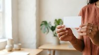 Woman with medium skin tone holding blank white card in hands, wearing rust-colored shirt indoors with plants and natural light for business, marketing, or greeting card mockup purposes.