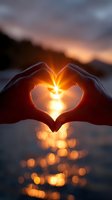 Hands forming heart shape against sunset sky with sun rays shining through fingers, creating golden bokeh reflection on water surface at dusk.