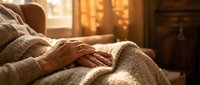 Elderly hands resting gently on a knitted blanket in a warm sunlit room, evoking comfort, care, and peaceful senior living at home.