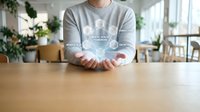 Person holding holographic cloud computing network icons with connected data symbols floating above hands in modern office workspace.