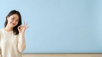 Young Asian woman in cream sweater waving hand against blue wall background, friendly greeting gesture for advertising, marketing, and web design projects.