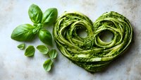 Heart-shaped homemade pasta with pesto sauce garnished with fresh basil leaves on textured stone background, perfect for Italian cuisine presentations.