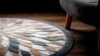 Circular mosaic rug with geometric leaf pattern in neutral beige and gray tones on dark wooden floor, modern home decor detail with partial armchair visible.