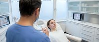 Young woman patient relaxing in modern dental chair during consultation with male dentist in bright clinical office with advanced equipment.