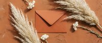 Elegant terracotta envelope surrounded by dried pampas grass and white flowers on textured clay surface for wedding invitations and stationery design.