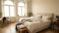 Serene bedroom with unmade bed, cream linens, arched windows, wooden floor, potted plants, and natural daylight for home decor and interior design projects.
