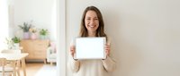 Woman with long brown hair, Caucasian ethnicity, wearing beige sweater, smiling while holding blank tablet with white screen for mockup design, modern home interior background.