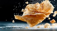 Crispy golden pastry shattering in motion with sugar powder splash against dark background, dynamic food photography capturing texture and movement.