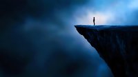 Silhouette of person standing on cliff edge against dramatic blue sky and clouds, representing challenge, risk, achievement and solitude in nature.
