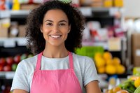 Young African American woman wearing pink apron smiling warmly in grocery store produce section, representing small business ownership and retail service.