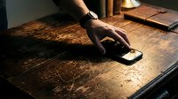 Male hand with dark skin tone reaching toward smartphone on wooden desk surface in warm lighting, suitable for technology, communication, and business concepts.