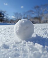 Round snowball on fresh white snow against blue winter sky background, natural sunlight creates soft shadows on crystalline surface. Perfect for winter season designs.