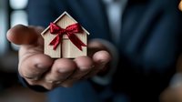 Small wooden house model with red festive bow held in male hand against dark background, symbolizing real estate, home ownership, and holiday season gifting.