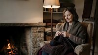 Middle-aged woman with dark hair and fair complexion, wearing a brown cardigan, sits in an upholstered armchair knitting by a fireplace with warm lighting and a floor lamp in a cozy home interior setting.