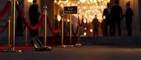 Elegant entrance with red velvet rope barriers, golden stanchions, and high heels on floor. Blurred guests in formal attire celebrate at luxury event with warm bokeh lights.