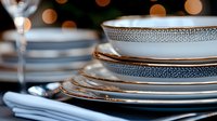 Stack of elegant porcelain plates with delicate silver trim pattern and wine glass against dark background with soft bokeh lights, luxury table setting.