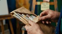 Senior man hands holding brown paper envelope filled with rolled dollar bills cash money in indoor setting with blurred background.