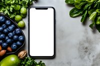 Smartphone mockup with blank white screen surrounded by fresh blueberries, limes, herbs and nuts on light gray concrete background. Food photography setup.