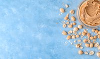 Creamy peanut butter in glass jar with scattered whole peanuts on blue textured background, top view with copy space for food photography and design.