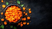 Fresh orange dried apricots in black ceramic bowl on dark stone background with scattered fruits and green leaves, top view with copy space.
