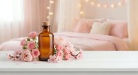 Amber glass bottle with pink roses on white surface in cozy bedroom with fairy lights. Spa wellness aromatherapy essential oil skincare beauty treatment concept.