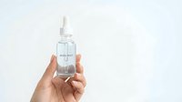 Clear glass dropper bottle with white cap held in hand against clean white background for skincare and cosmetic product marketing.