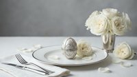 Elegant Easter table setting with decorative silver egg on white plate, cream roses and silverware for spring holiday dining and celebration.