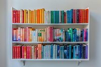 Colorful books arranged by rainbow spectrum on white floating shelves against light wall, creating vibrant home library display for interior design and education concepts.
