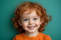 Happy toddler with curly red hair and bright smile wearing orange shirt against turquoise background for children photography and family portraits.