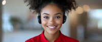 Young African American woman with natural curly hair wearing red sweater and wireless headphones smiles warmly at camera against soft bokeh background.