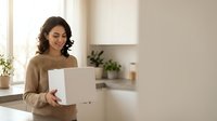 Happy young Hispanic woman holding white delivery box in modern kitchen, smiling while receiving online shopping package at home counter.