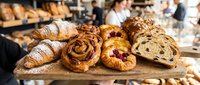 Freshly baked pastries on wooden board in artisan bakery, croissants, danish, cinnamon rolls displayed with blurred staff background.