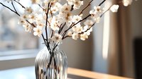 Delicate white cherry blossoms in striped glass vase on wooden table, soft natural window light creates dreamy atmosphere with blurred background.
