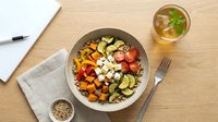 Healthy grain bowl with roasted vegetables, feta cheese, and fresh tomatoes on a wooden table with iced tea and notebook.