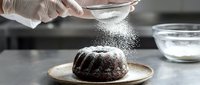 Gloved hand dusting powdered sugar onto freshly baked chocolate cake in professional kitchen setting for baking, pastry, and culinary content.