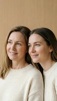 Two Caucasian women mother and daughter in beige sweaters embracing warmly against neutral background for family lifestyle content.