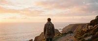 Hiker with backpack standing on coastal cliff overlooking ocean at sunset, adventure travel and outdoor exploration concept for lifestyle and wanderlust themes.